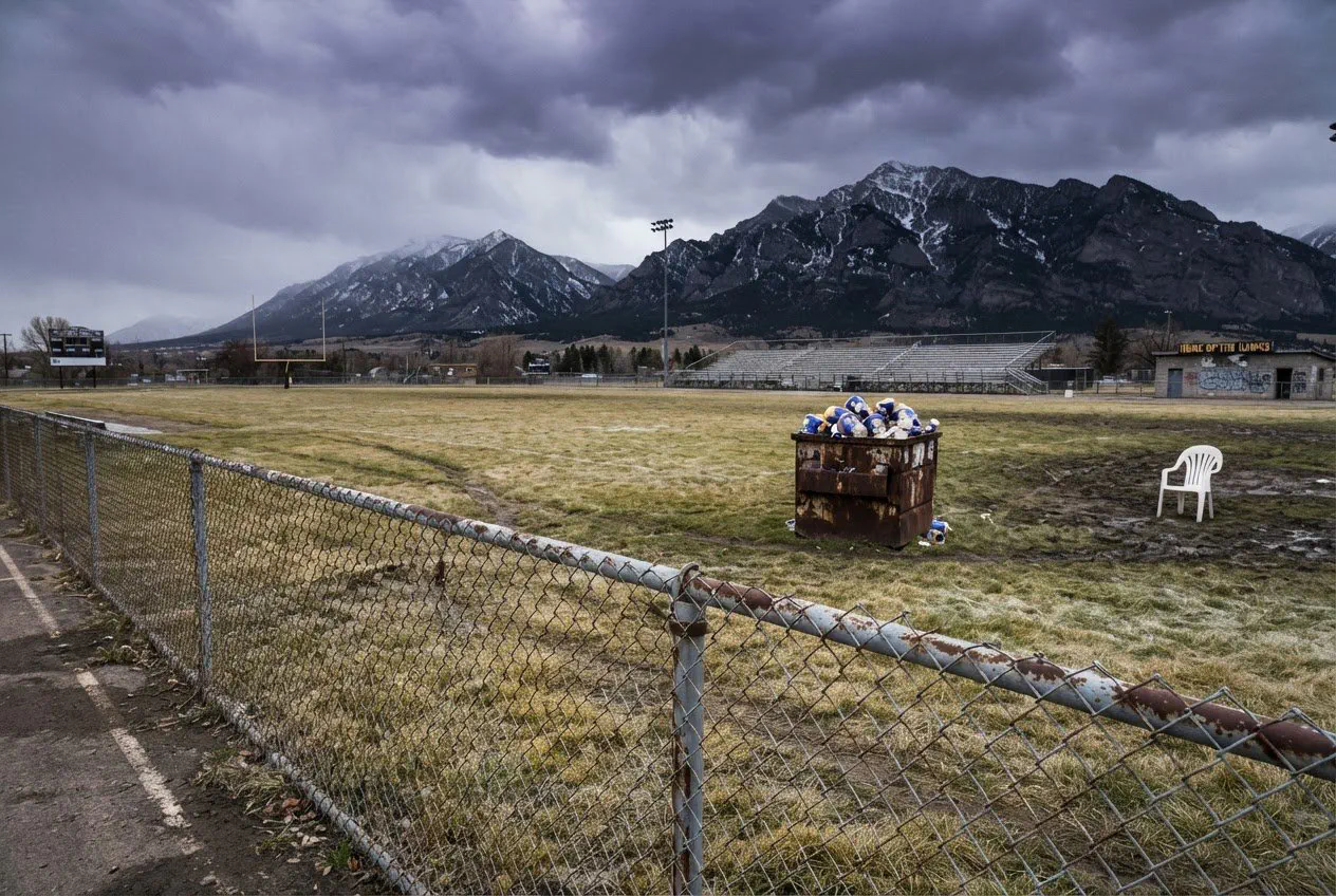 A wide, eye-level landscape shot of the empty grass field at Van Winkle Stadium in late autumn, looking toward the Bridger Range under a heavy, bruised-purple