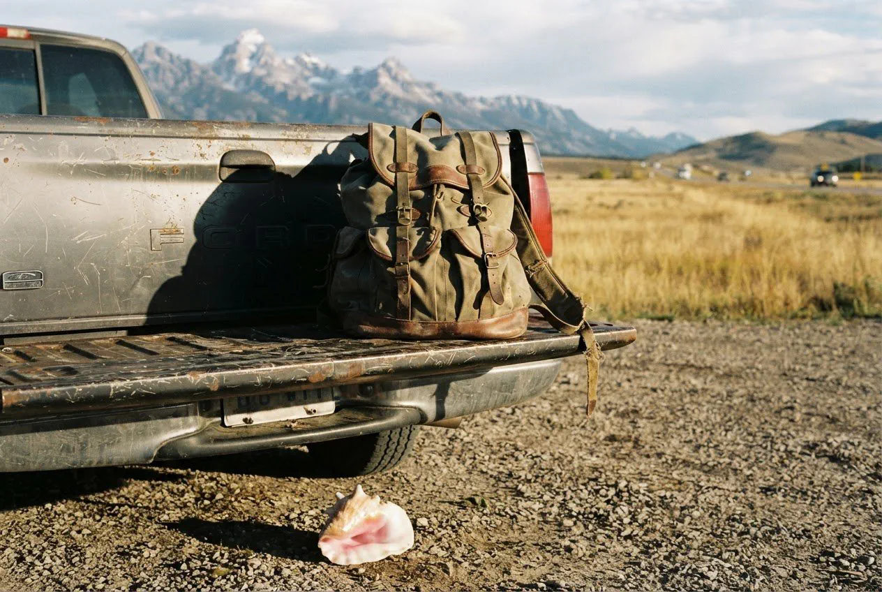 A close-up, eye-level shot of a weathered, canvas-and-leather rucksack sitting on the rusted, scratched metal tailgate of a 1998 Ford F-150.